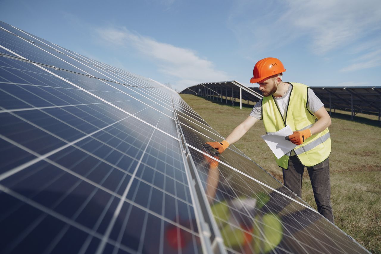 gallery-4 Young bearded male engineer in protective helmet and heat resistant gloves touching solar panel while standing on field with papers on sunny day